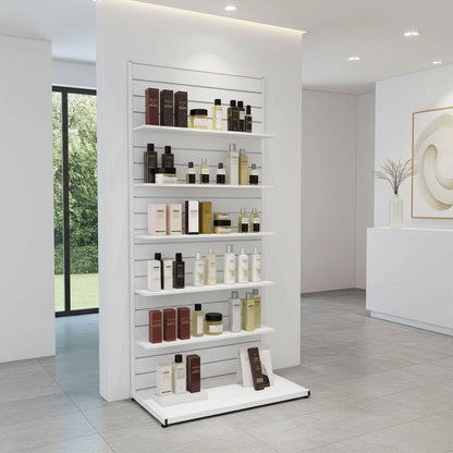 A white slatwall display shelf unit in a modern beauty salon, stocked with various skincare and cosmetic bottles and jars. The room features light gray tiled floors, white walls, and a large window on the left showing greenery outside.