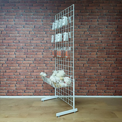 A white metal wire grid display stand with a lower wire basket, shown against a red brick wall and light wood floor. Several small white items are hanging on the grid, and a basket of white wool.